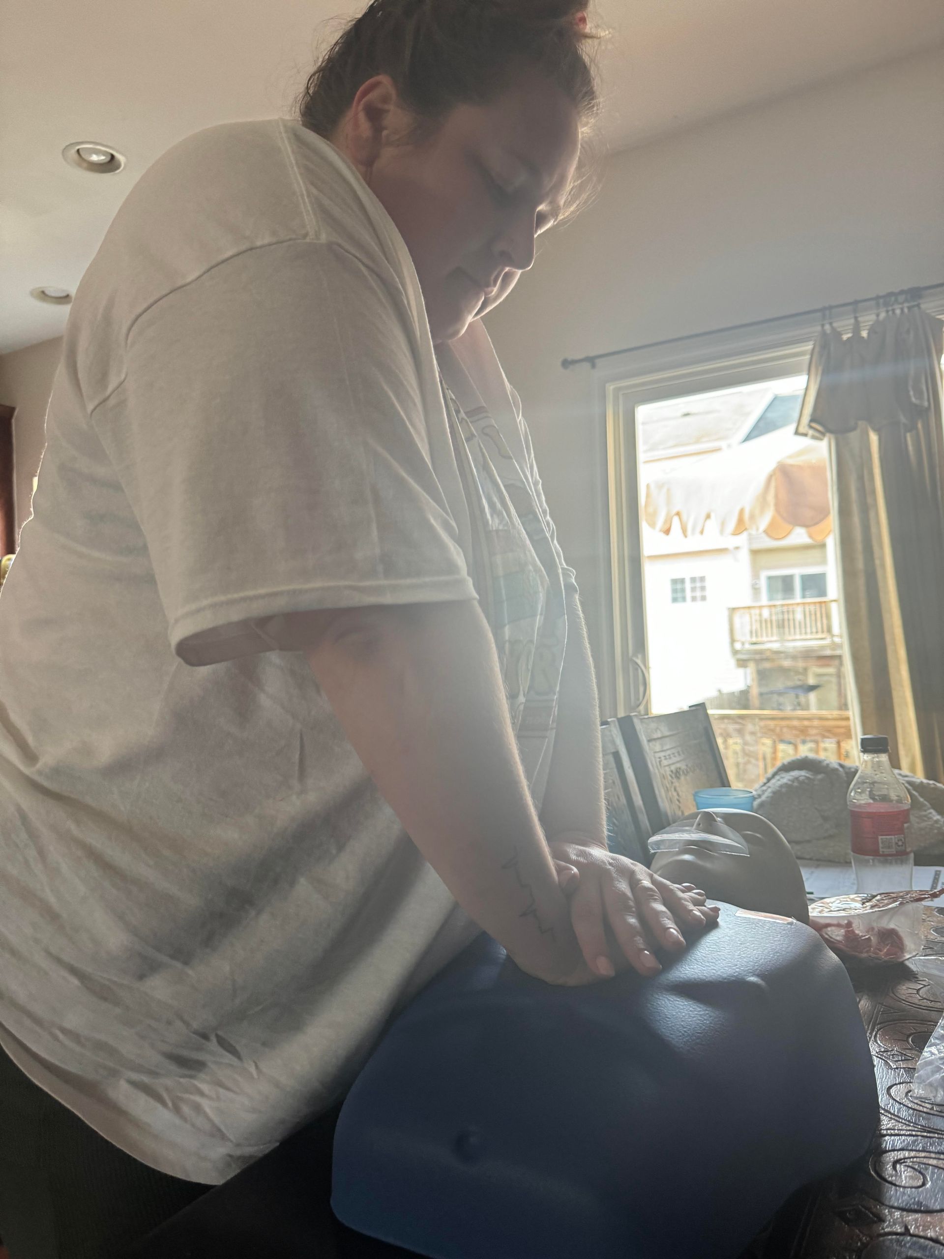 Person in a white shirt performing CPR on a blue dummy indoors.