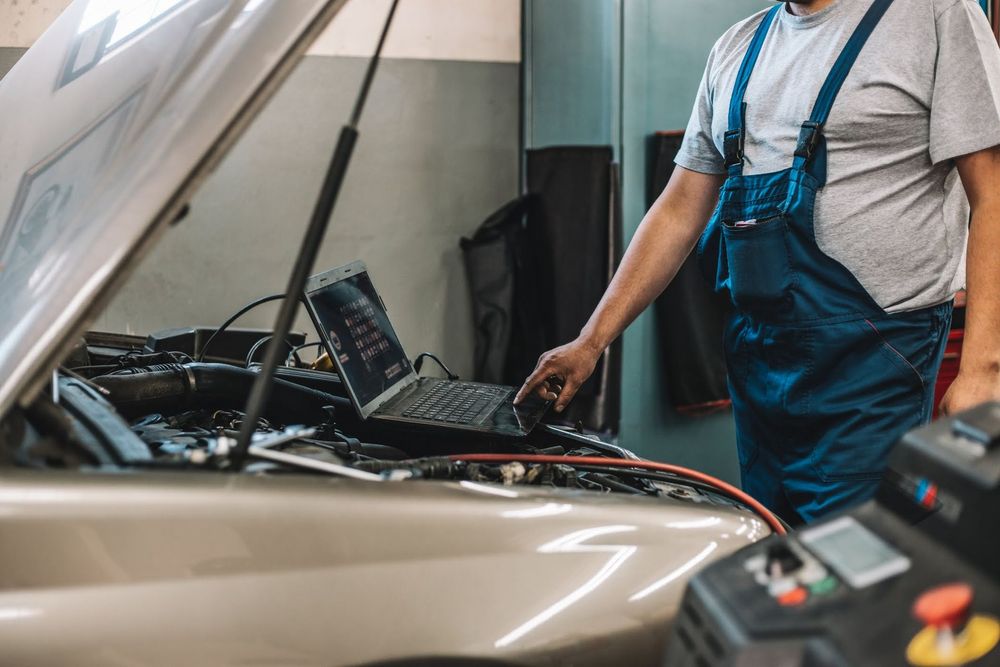 Un meccanico in tuta blu controlla il vano motore con il cofano aperto in un'officina.