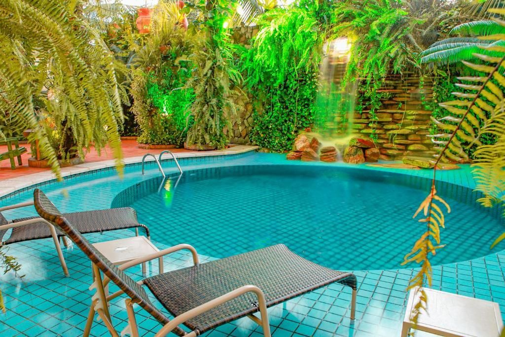 Uma piscina no quintal rodeada por vegetação exuberante e uma pequena cascata, com duas espreguiçadeiras na lateral.