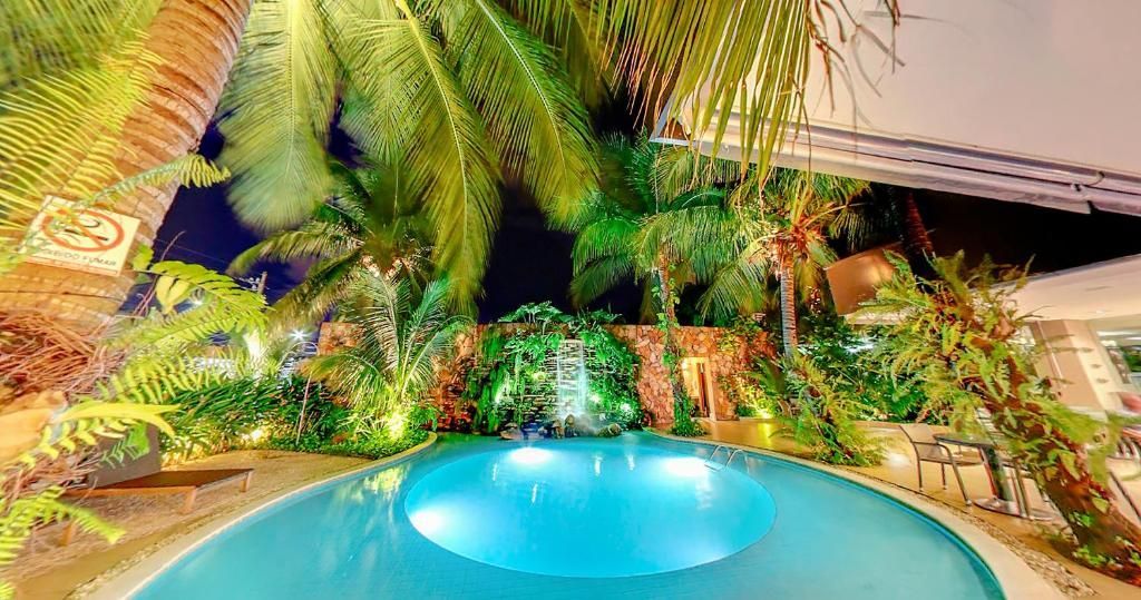 Piscina rodeada por plantas tropicais e uma pequena cascata à noite.