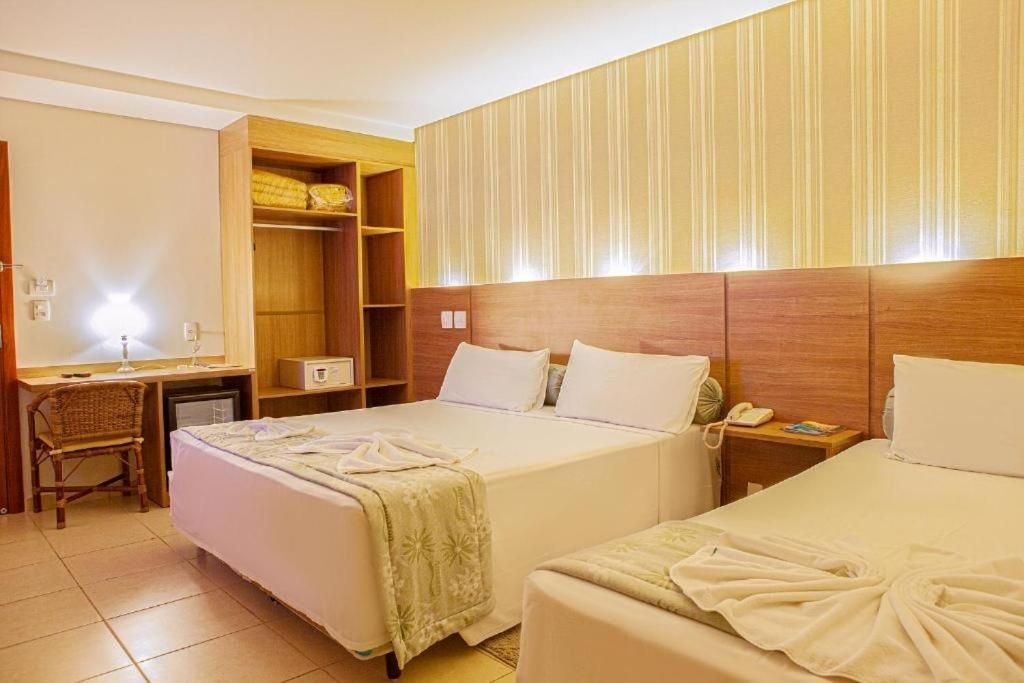 Quarto de hotel com duas camas, guarda-roupa de madeira, escrivaninha e paredes em tons neutros.