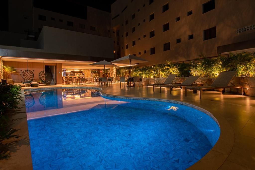Vista noturna de uma piscina com um prédio e um pátio ao fundo; iluminação quente.