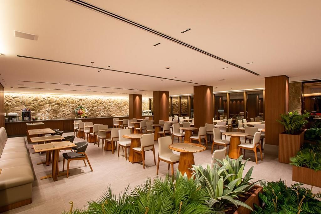 Área de refeições do restaurante, esquema de cores neutras, mesas e cadeiras dispostas, plantas, iluminação suspensa.