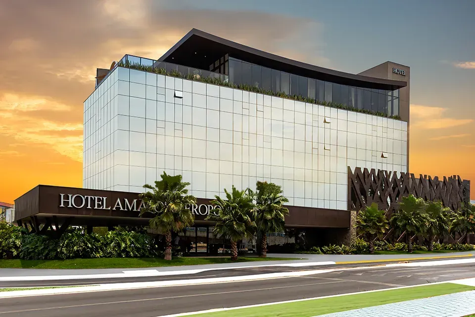 Prédio de hotel moderno com fachada de vidro e sinalização; palmeiras e rua em primeiro plano.