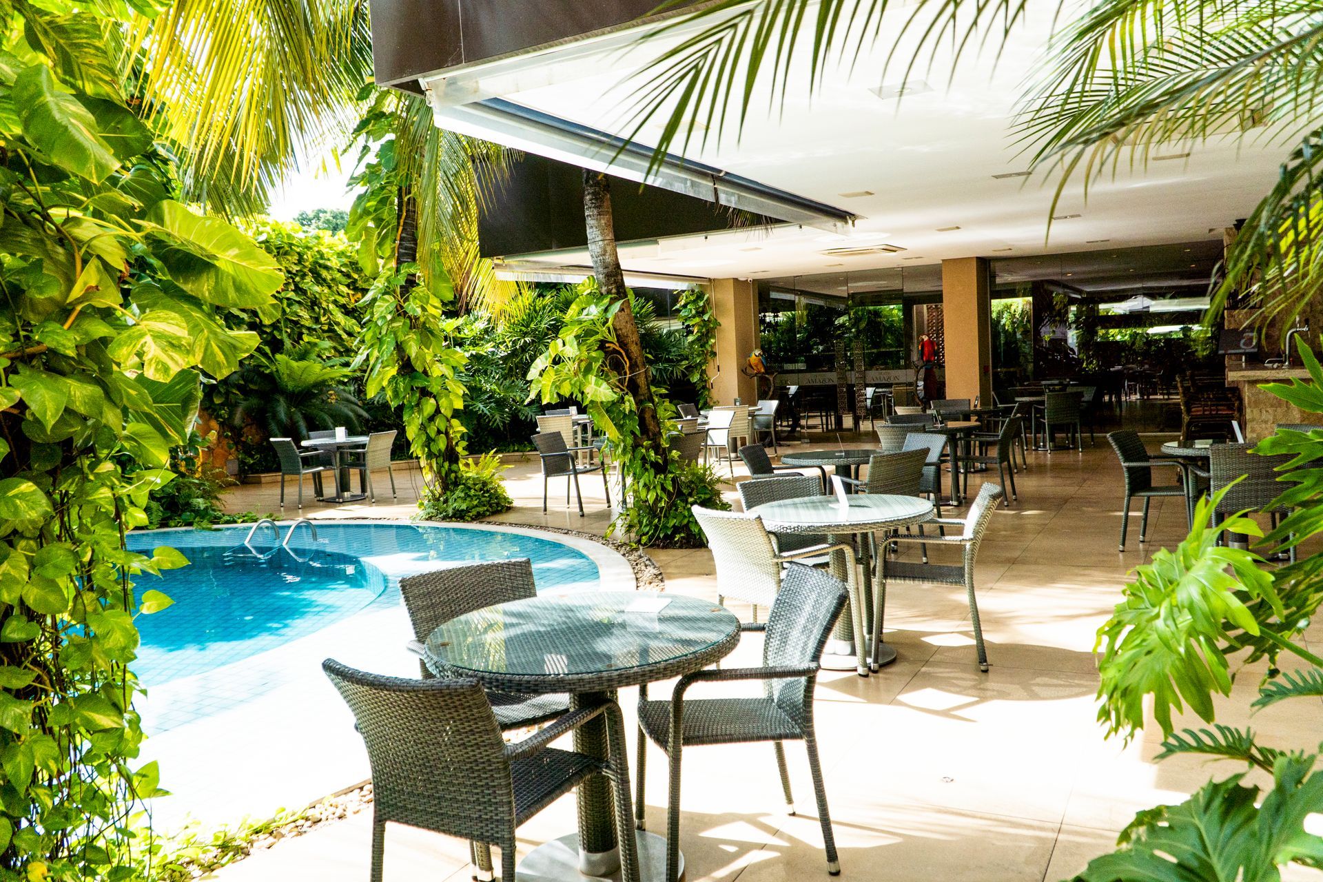 Área de jantar ao ar livre com mesas, cadeiras e piscina. O espaço é rodeado por uma vegetação exuberante.