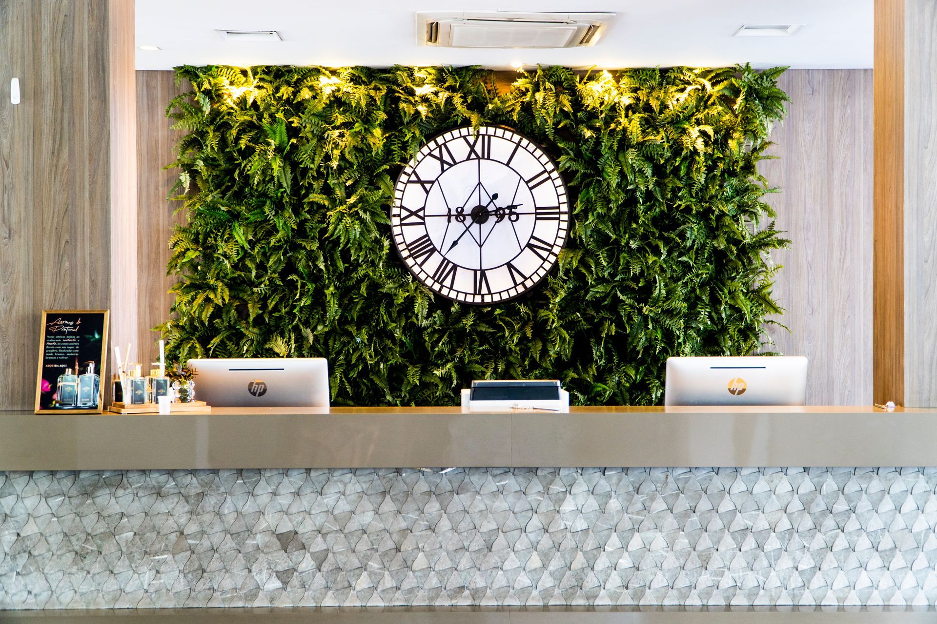 Balcão de recepção do hotel com parede verde, relógio e computadores.