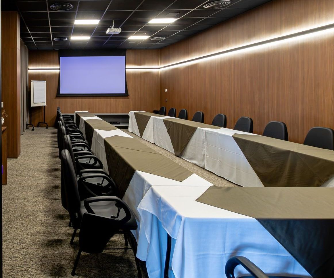 Sala de conferências com mesa em formato de U, toalhas de mesa brancas e cinzas, cadeiras pretas e um biombo.