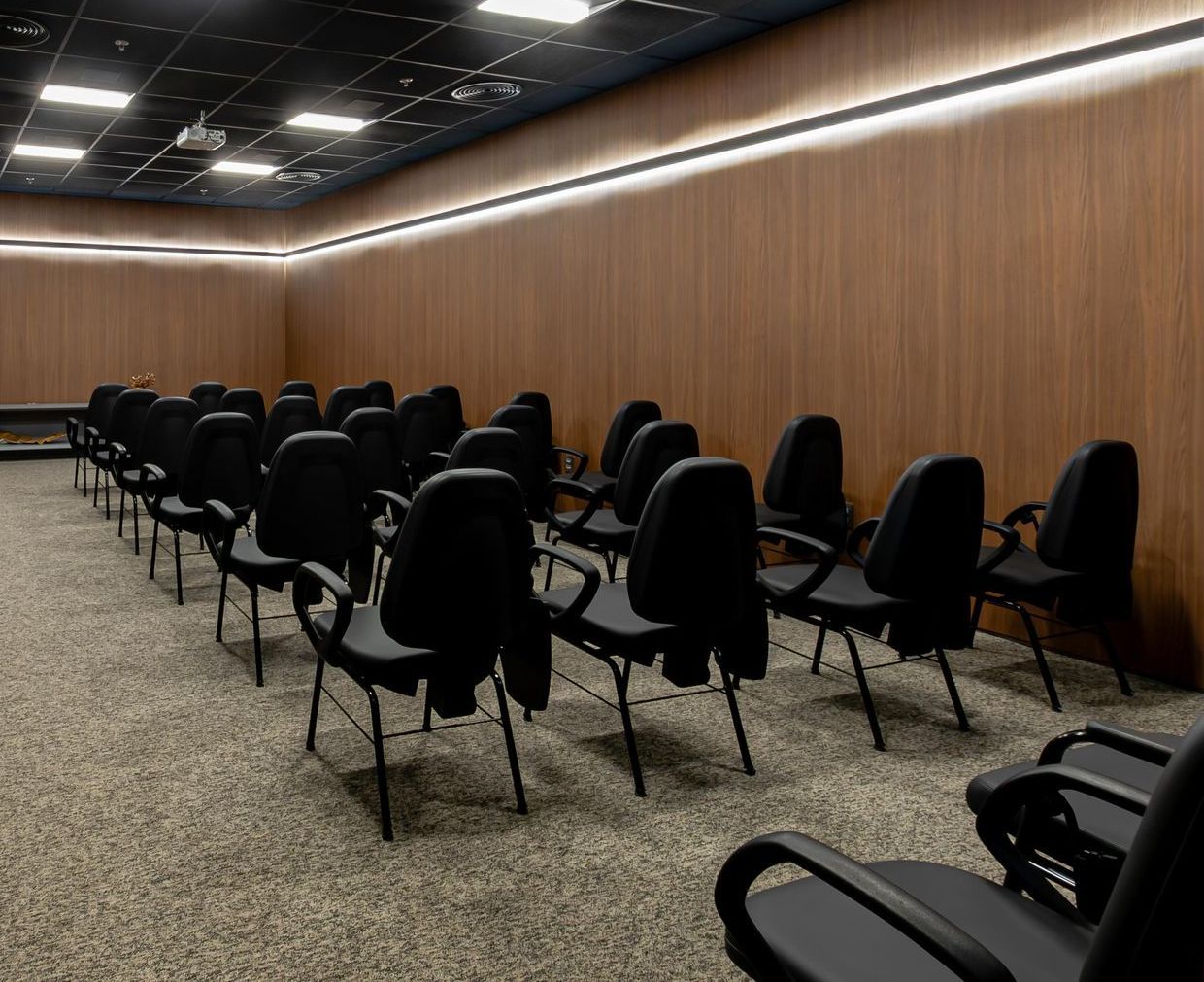 Fileiras de cadeiras pretas em uma sala de conferências vazia, com painéis de madeira e piso acarpetado.