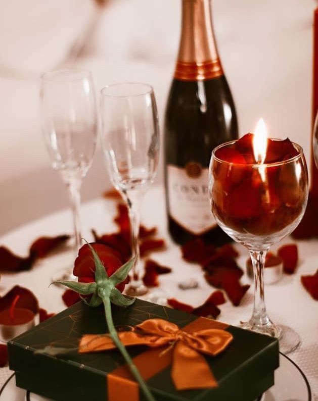 Mesa posta de forma romântica: champanhe, vela acesa, caixa de presente com uma rosa e pétalas espalhadas.