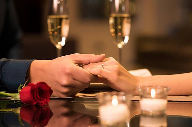 Mãos entrelaçadas sobre uma mesa com velas, rosas vermelhas e taças de champanhe, sugerindo um jantar romântico.