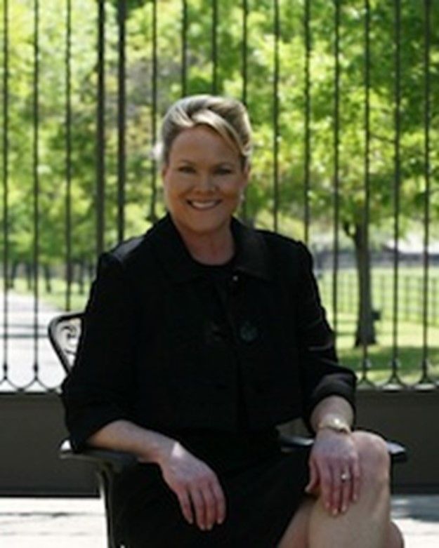 Woman in black suit smiles, sitting outside, black gate in background.