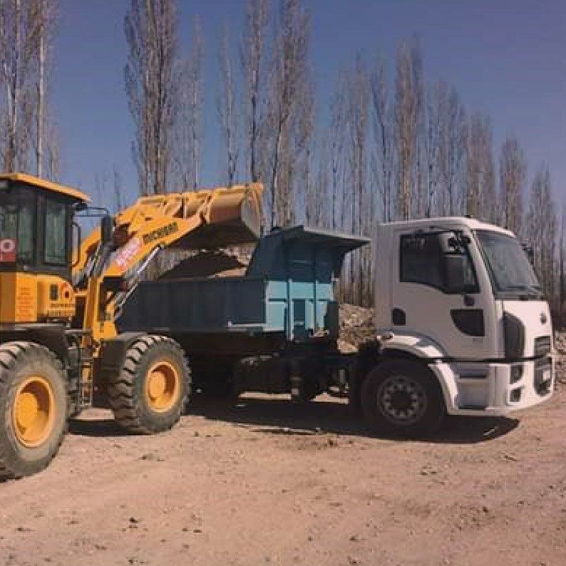 Una cargadora amarilla descarga material en un camión azul junto a una cabina blanca en un camino de tierra.