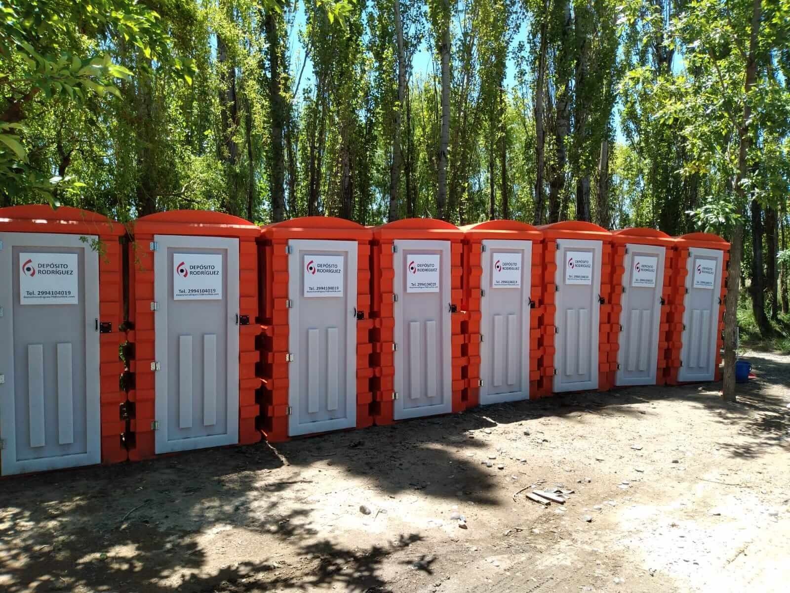 Hilera de baños portátiles grises con tapas rojas en una zona boscosa al aire libre.