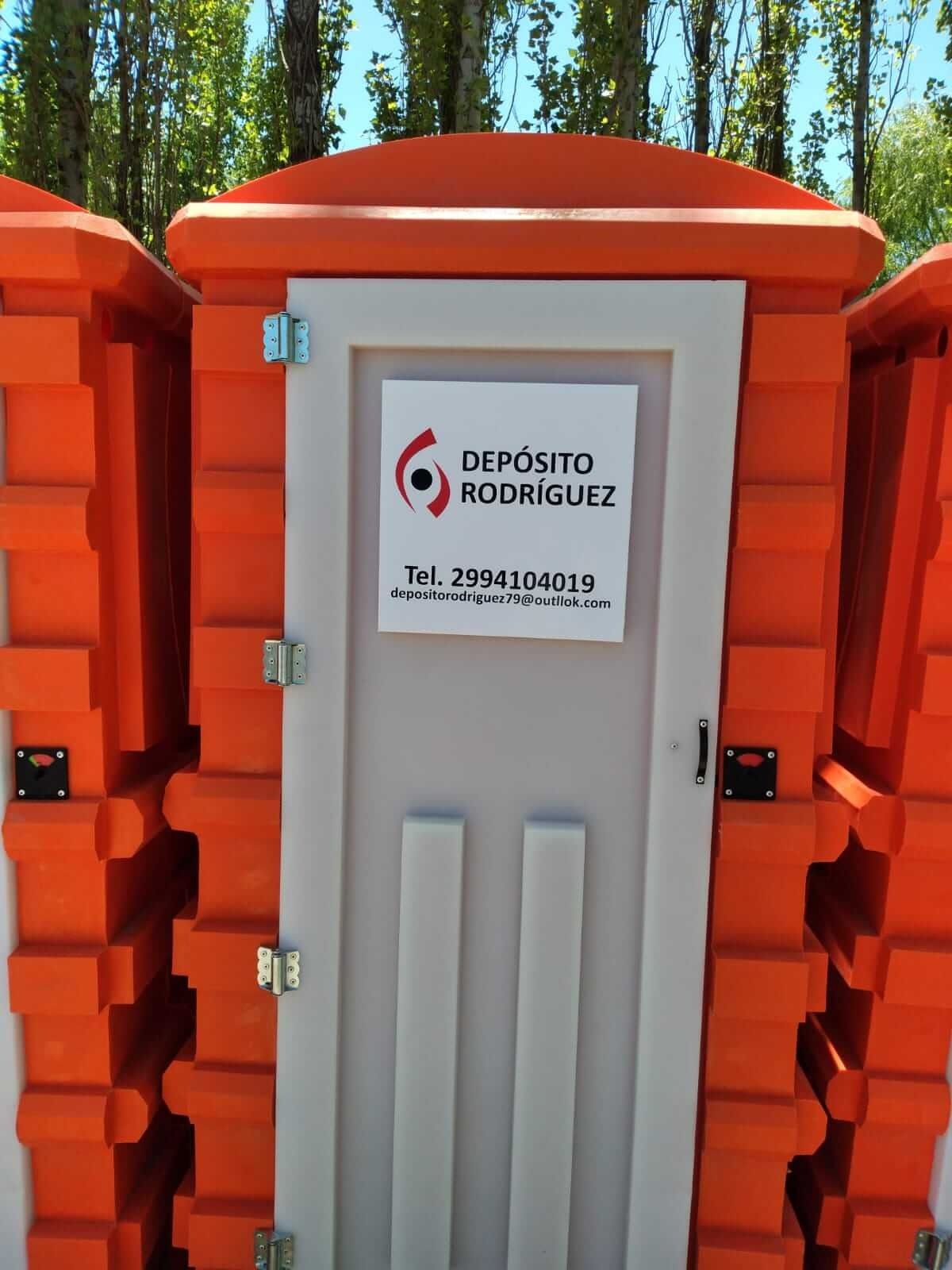 Baño portátil gris con letrero “Depósito Rodríguez”, rodeado de barreras naranjas en el exterior.