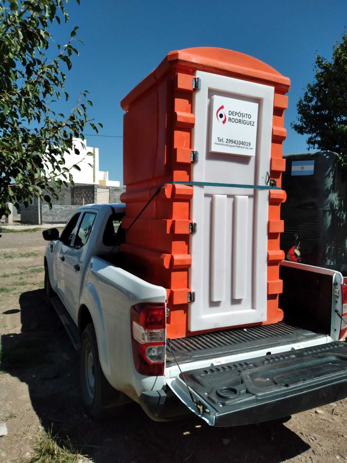 Un inodoro portátil naranja sujeto a la caja de una camioneta blanca al aire libre.