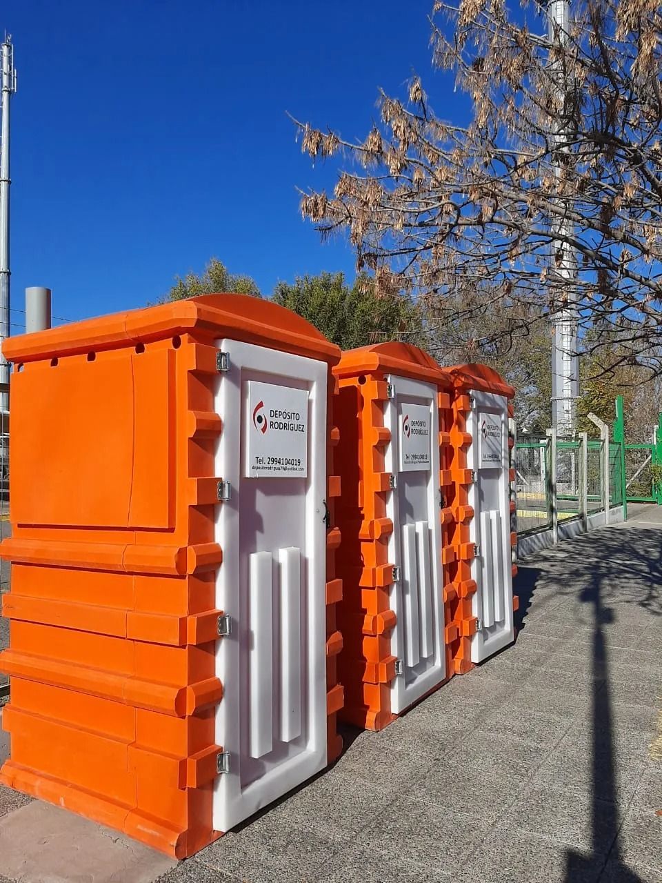 Baños portátiles naranjas alineados al aire libre en un día soleado con cielo azul y árboles cerca.