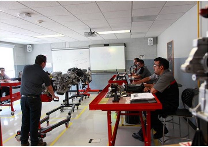 Sala de aula de mecânica automotiva. Instrutor com motor, alunos em mesas vermelhas com laptops, quadro branco.