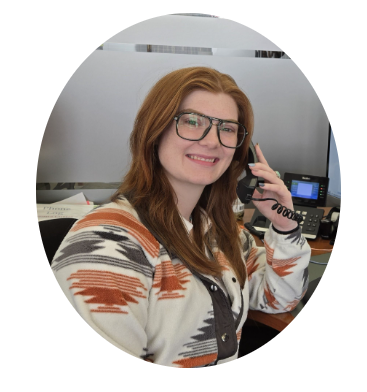A person with long red hair and glasses smiles while holding a telephone handset in an office setting.