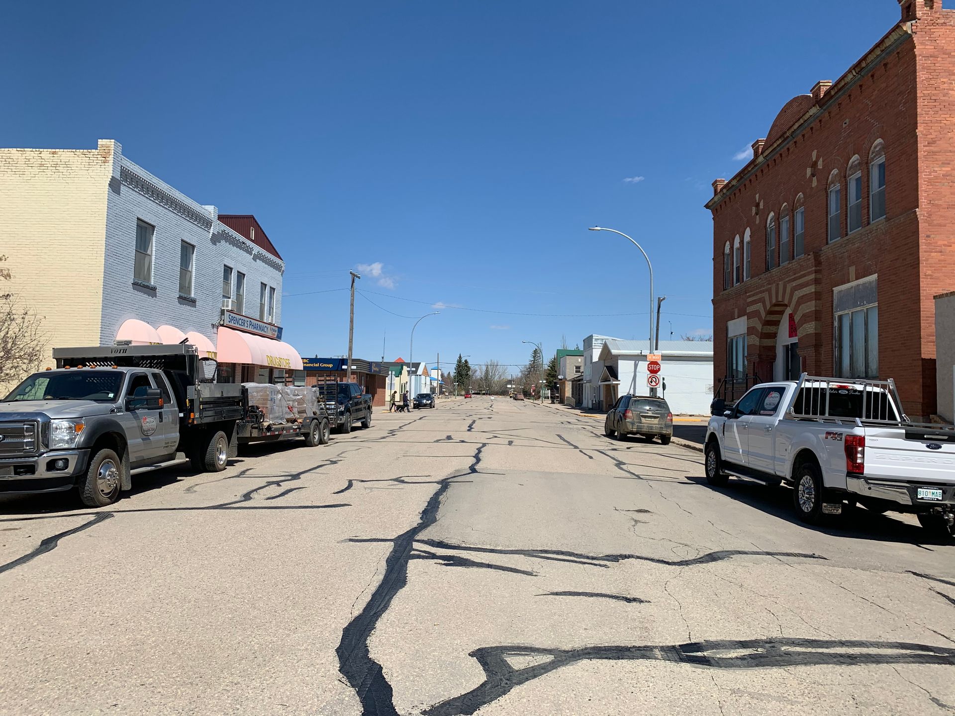 A quiet, sunny street in a small town with historic buildings, parked trucks, and a cracked asphalt road.