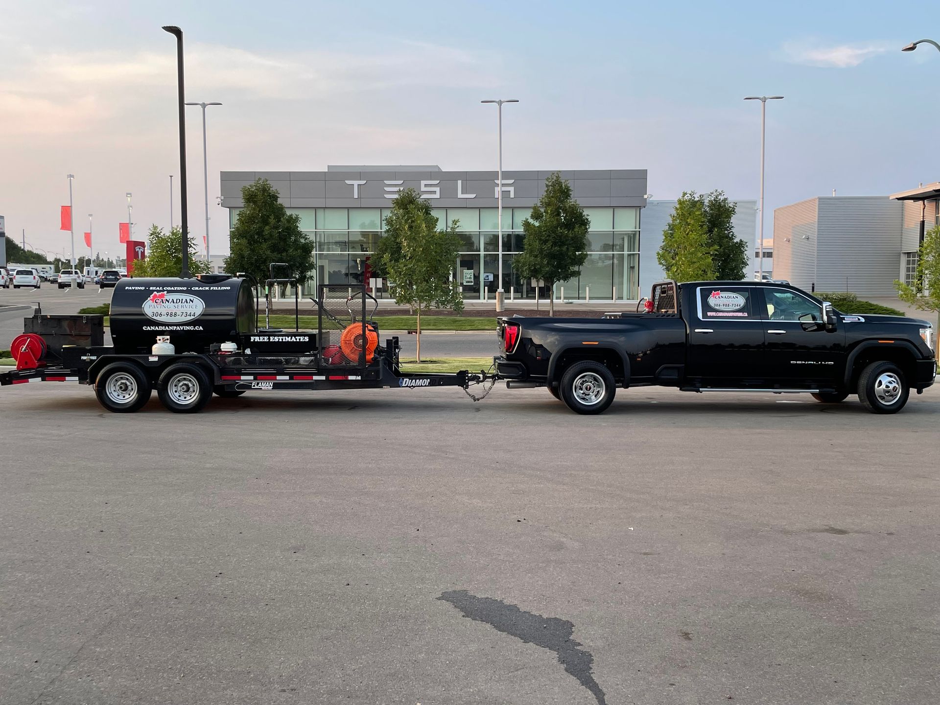 A black pickup truck towing a fuel tank trailer parked in front of a Tesla dealership.