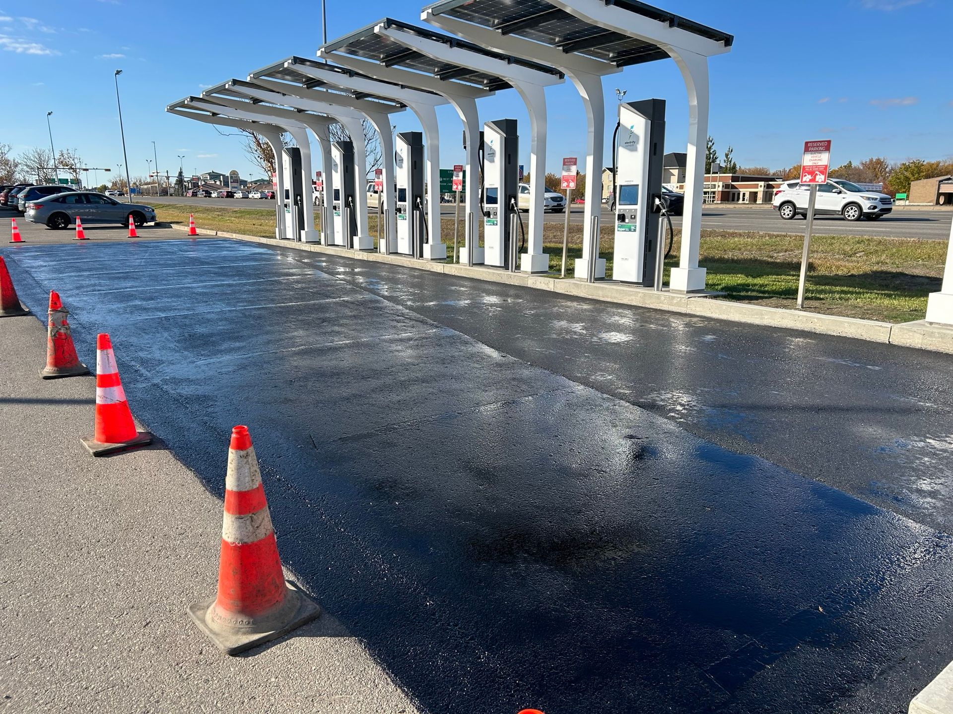 A row of electric vehicle charging stations under a solar canopy with a freshly paved, wet asphalt surface and traffic cones.