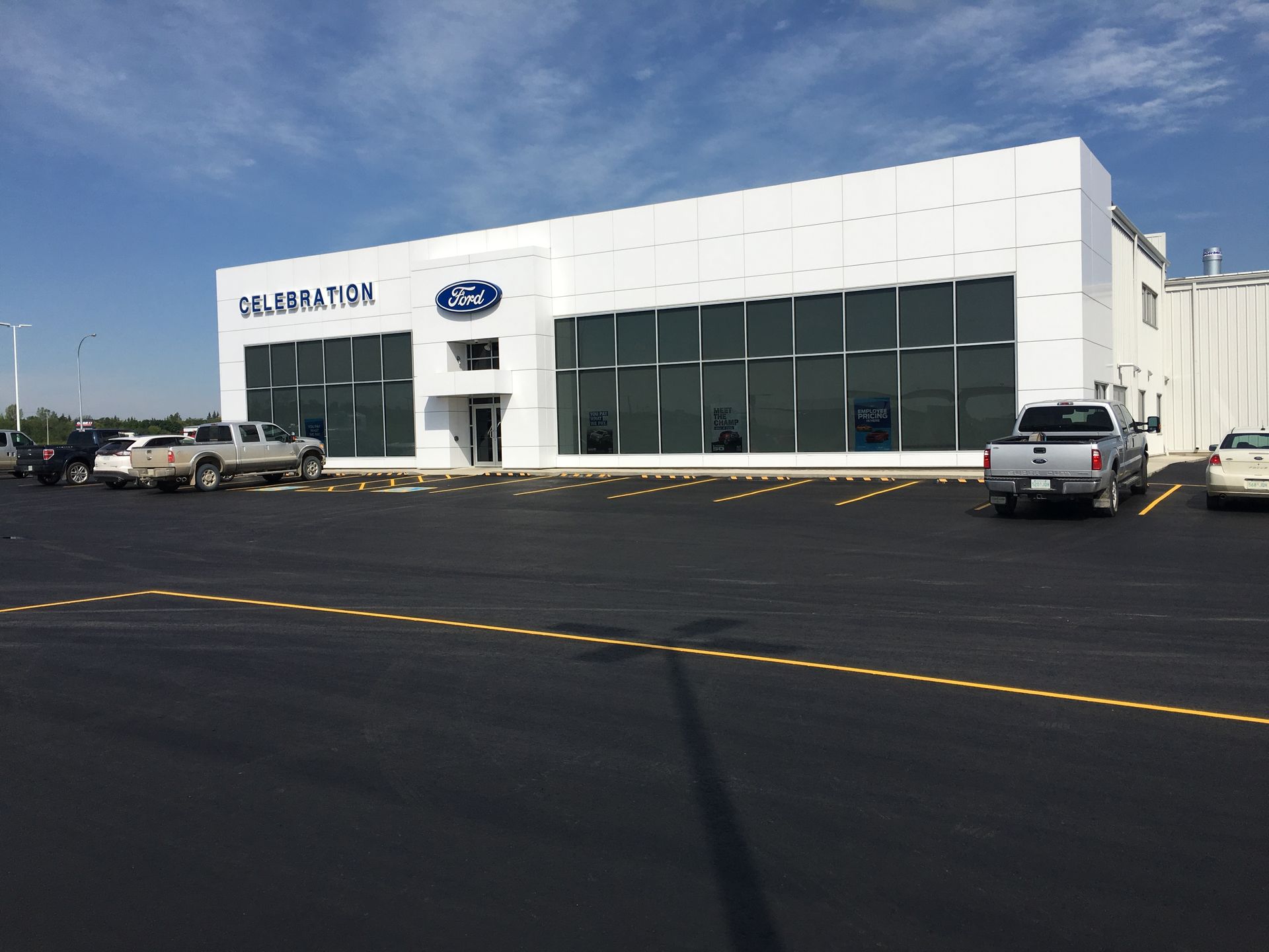 A white Ford dealership building with large glass windows and parked cars on an asphalt lot under a blue sky.