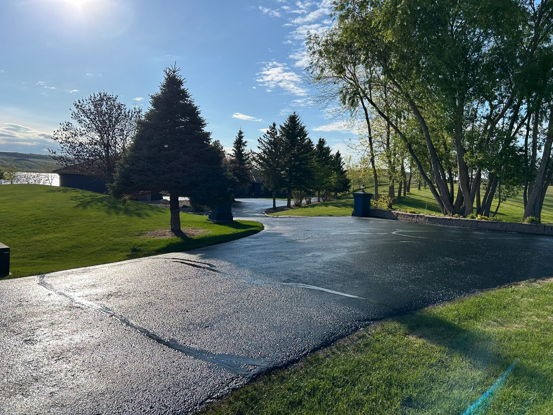 A newly paved asphalt driveway curves past a green lawn and evergreen trees under a clear, sunny sky.