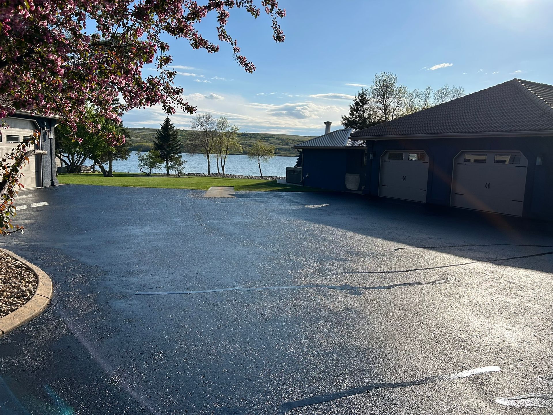 A freshly sealed asphalt driveway leads toward a garage and a scenic view of a lake under a bright, sunny sky.