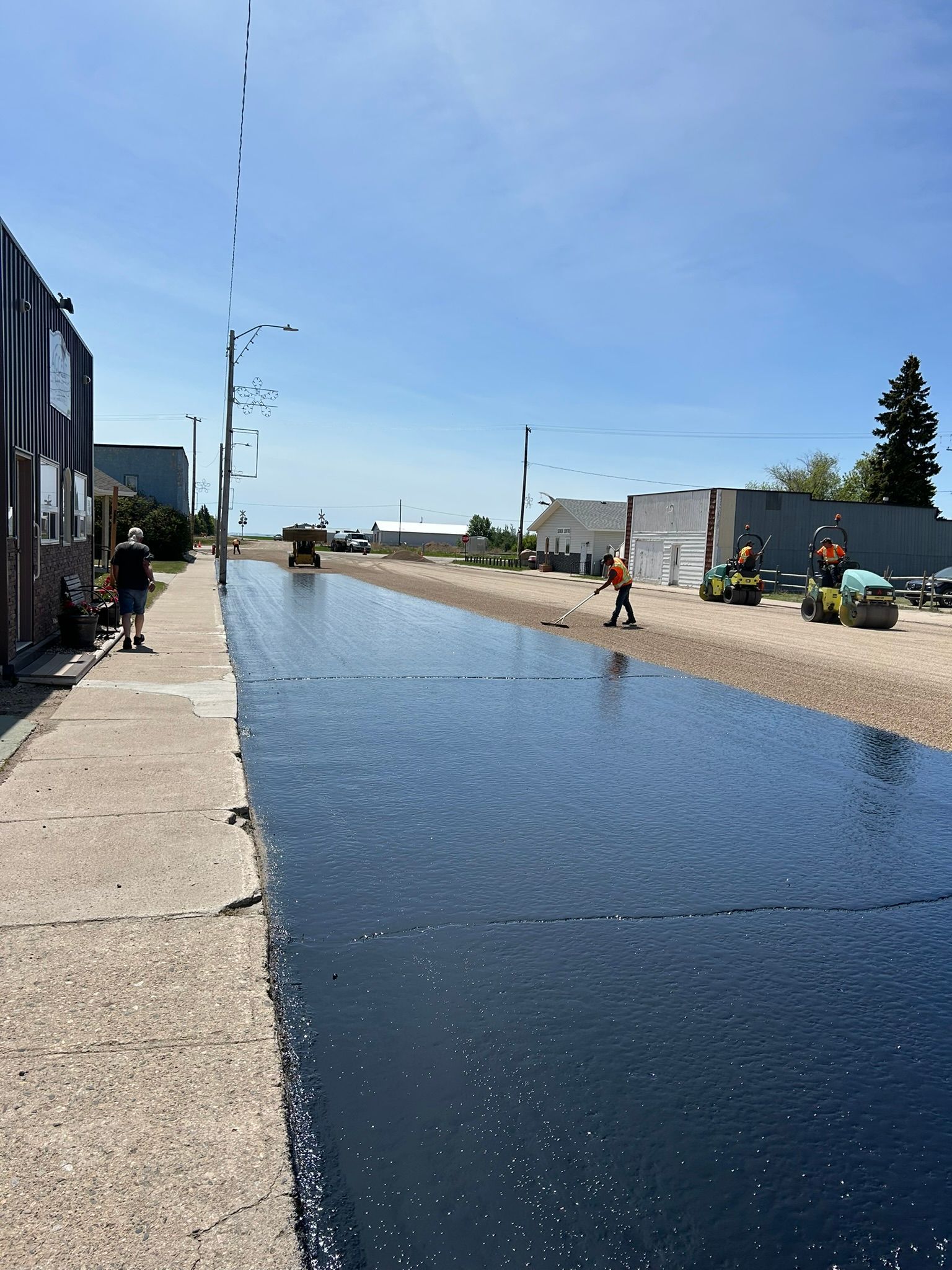 Construction workers apply a black adhesive coat to a road surface in a sunny, urban environment.