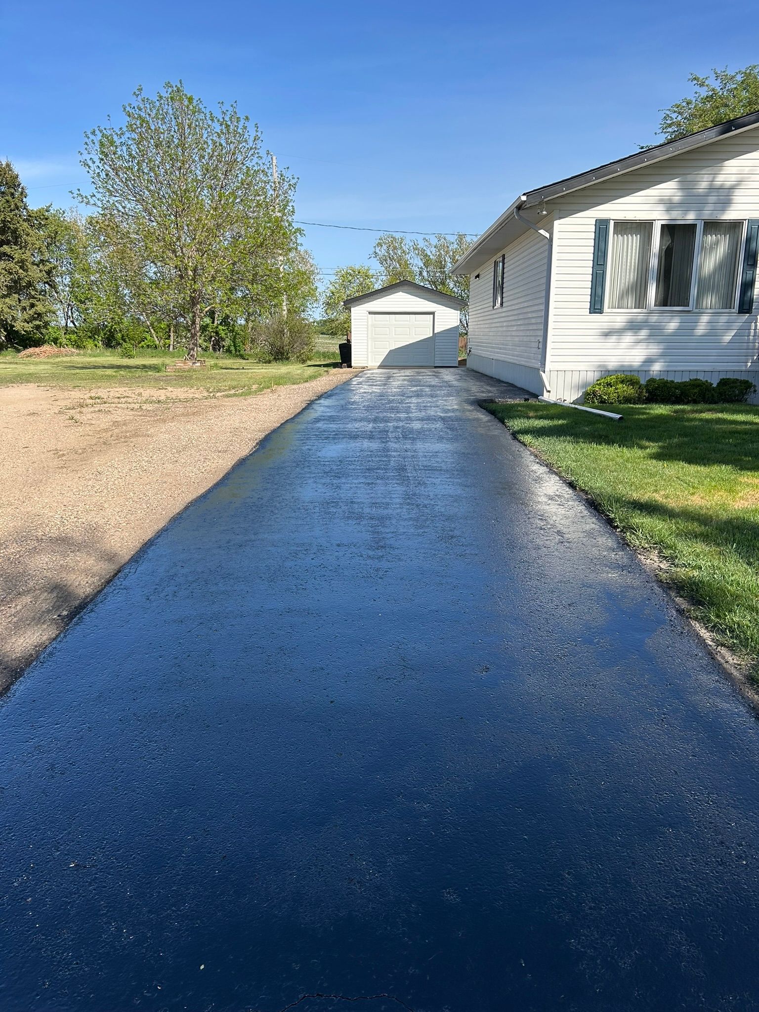 A freshly seal-coated, dark black asphalt driveway leading to a garage next to a white house with blue shutters.