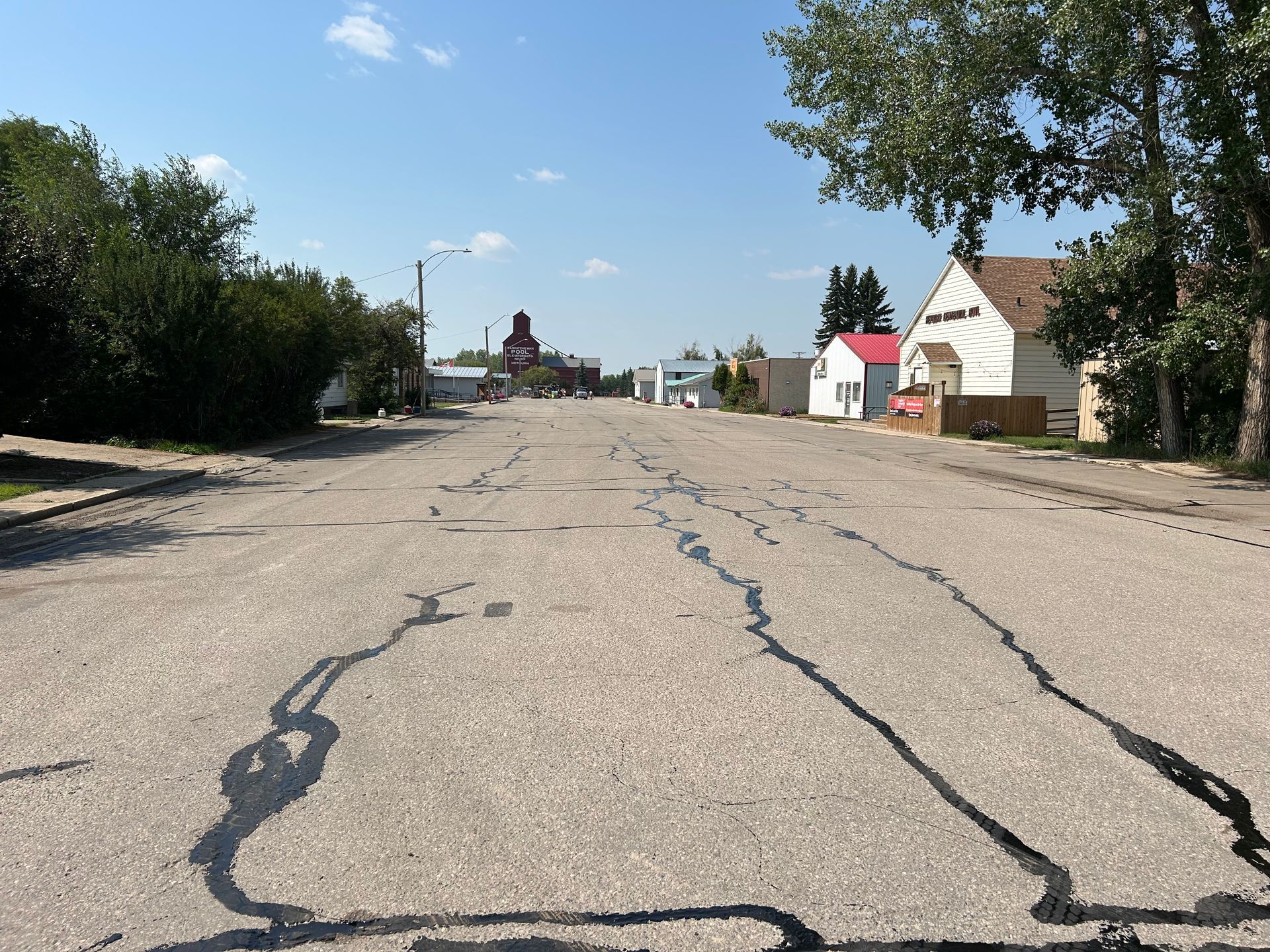 A paved, cracked road stretches through a small, quiet town with buildings on the sides under a clear blue sky.