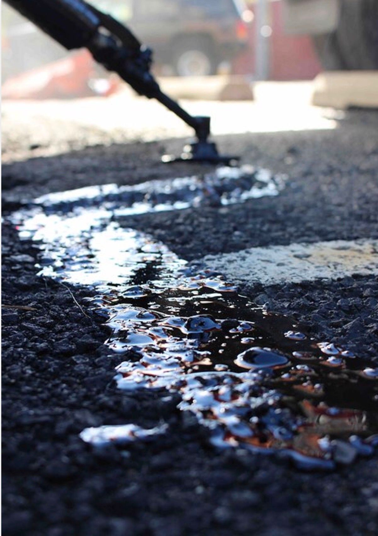 A close-up of a heated crack-sealing wand applying molten black asphalt filler to a crack in a dark paved road.