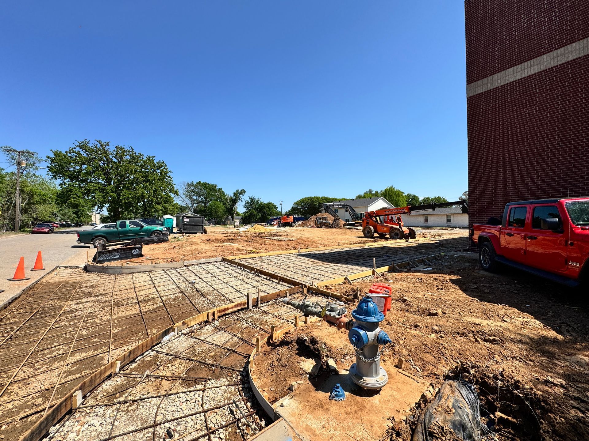 Construction site with exposed rebar, concrete forms, and equipment. A red building and truck are visible. Bright blue sky.