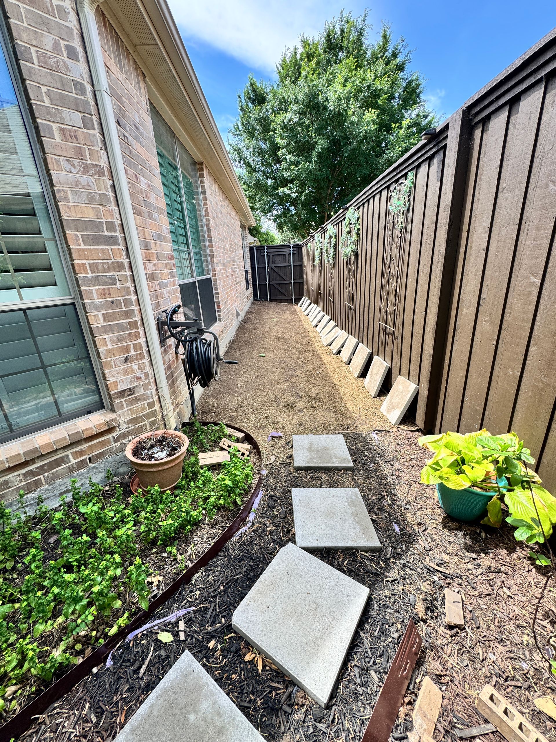 Narrow yard with brick wall, brown fence, stepping stones, and mulch pathway.