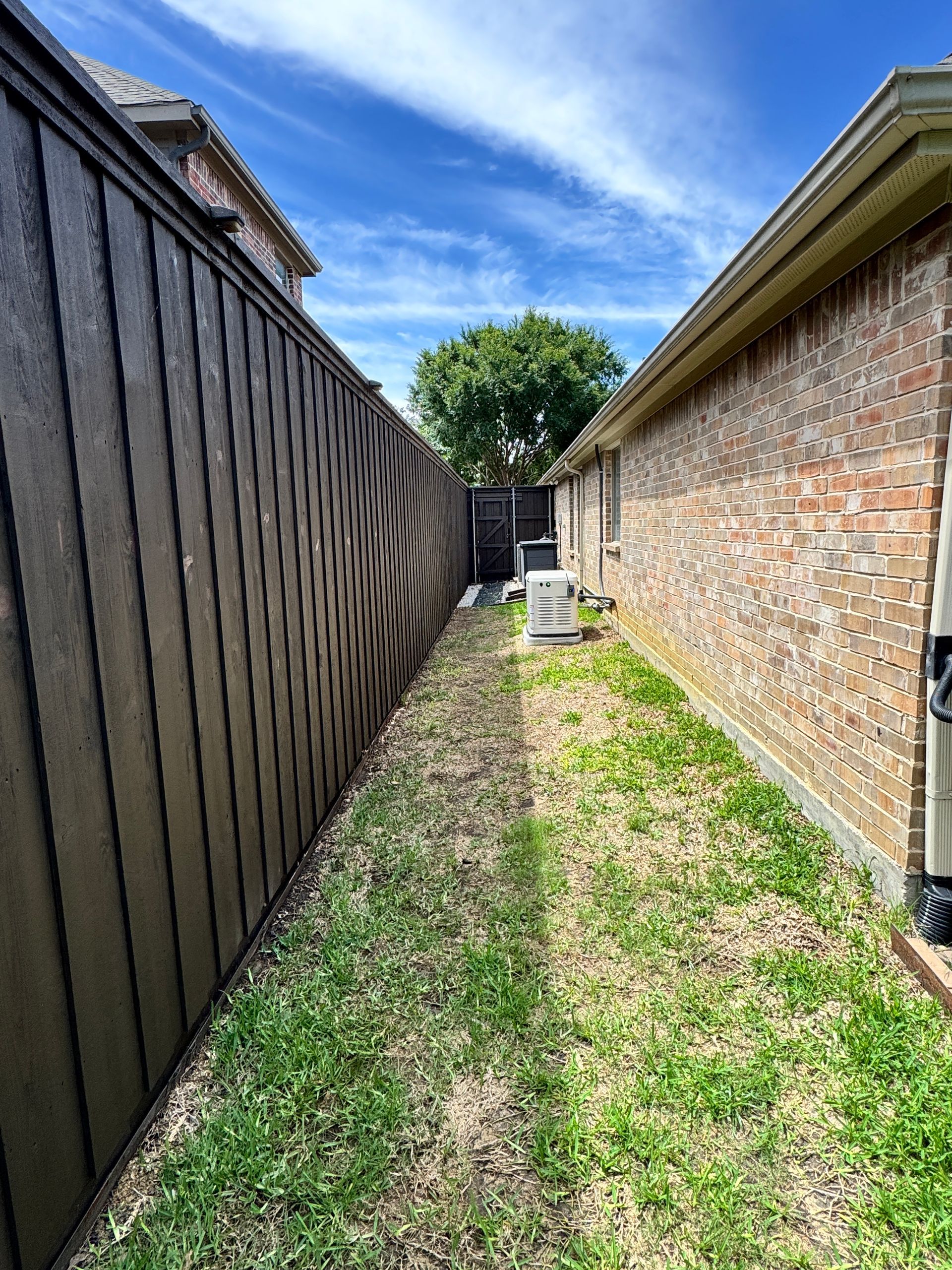 Narrow yard between a brown fence and brick house, with grass, air conditioning units, and blue sky.