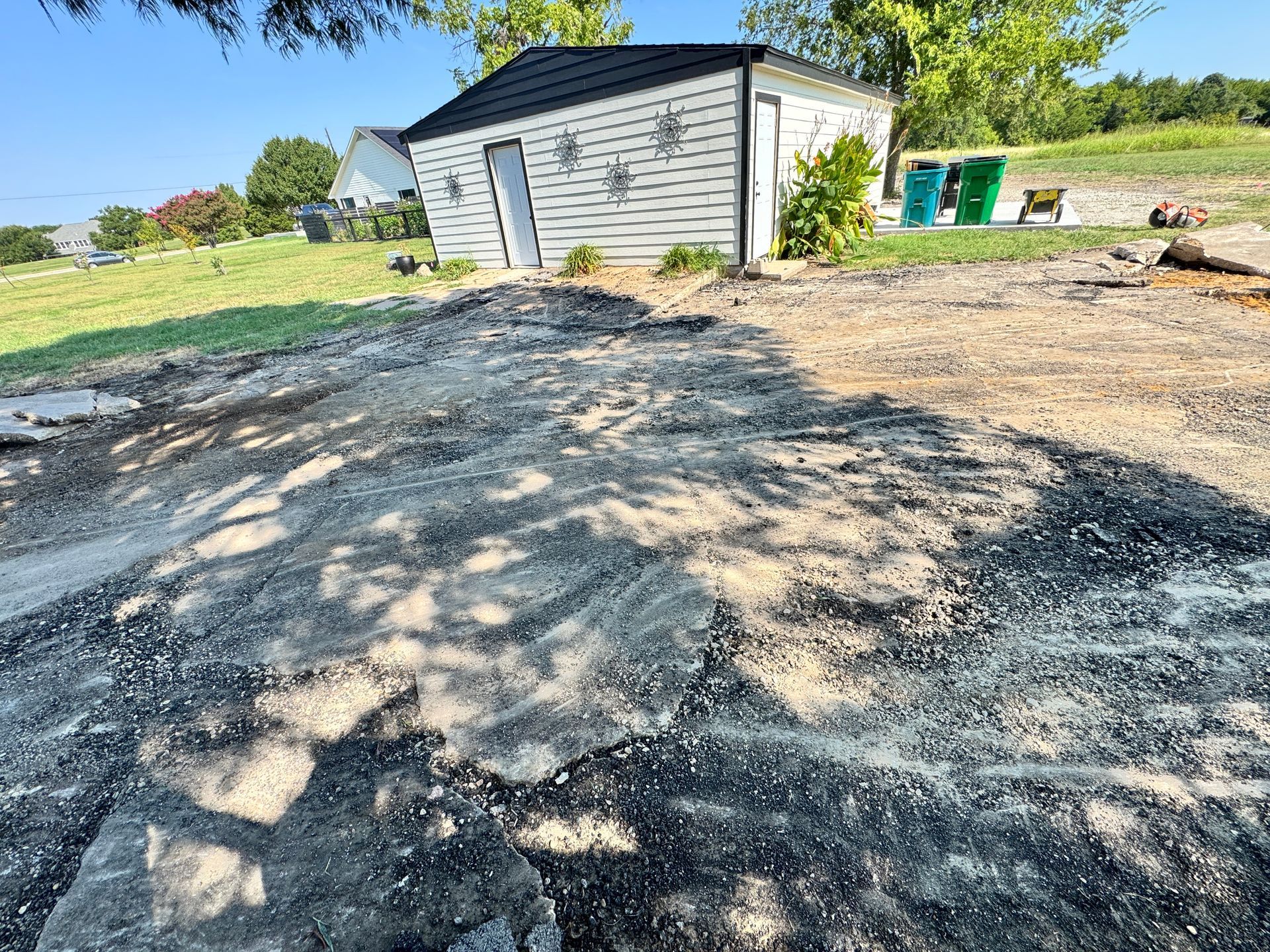 Gravel area in front of a small white building with black roof and green trash cans on grass, sunny day.