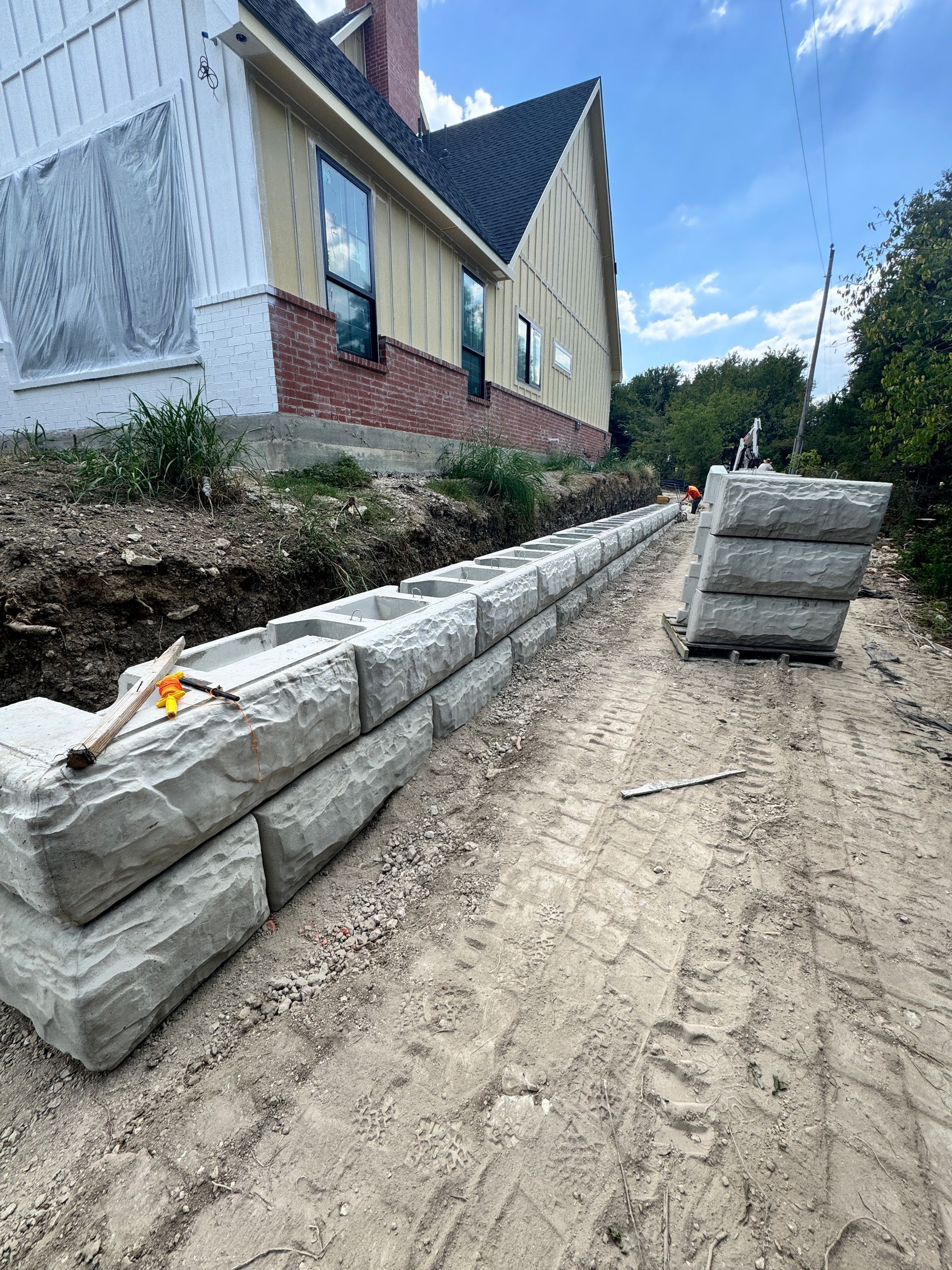 Retaining wall under construction next to a house with exposed siding. Gray stone blocks are stacked.