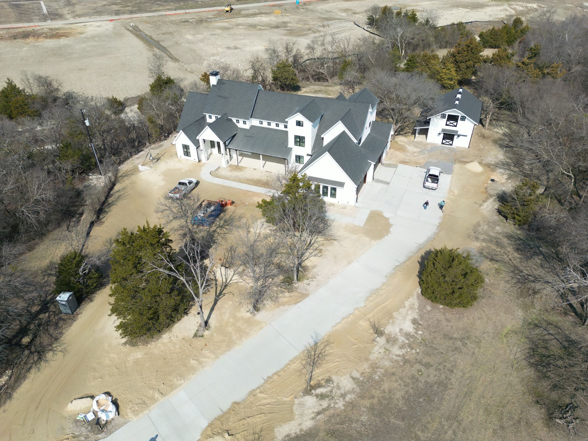 Aerial view of a white modern house with a dark roof on a hill, with a long driveway and bare trees.