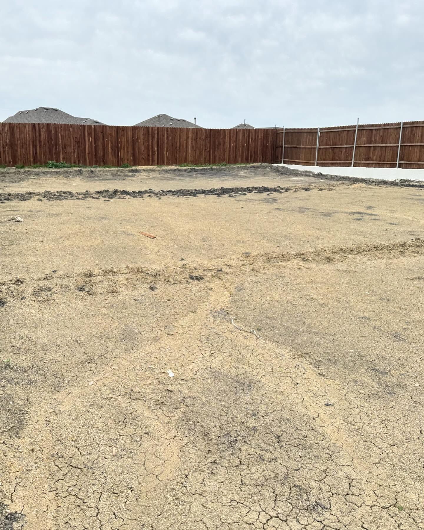 Dirt yard with tire tracks, enclosed by a brown wooden fence under a cloudy sky.