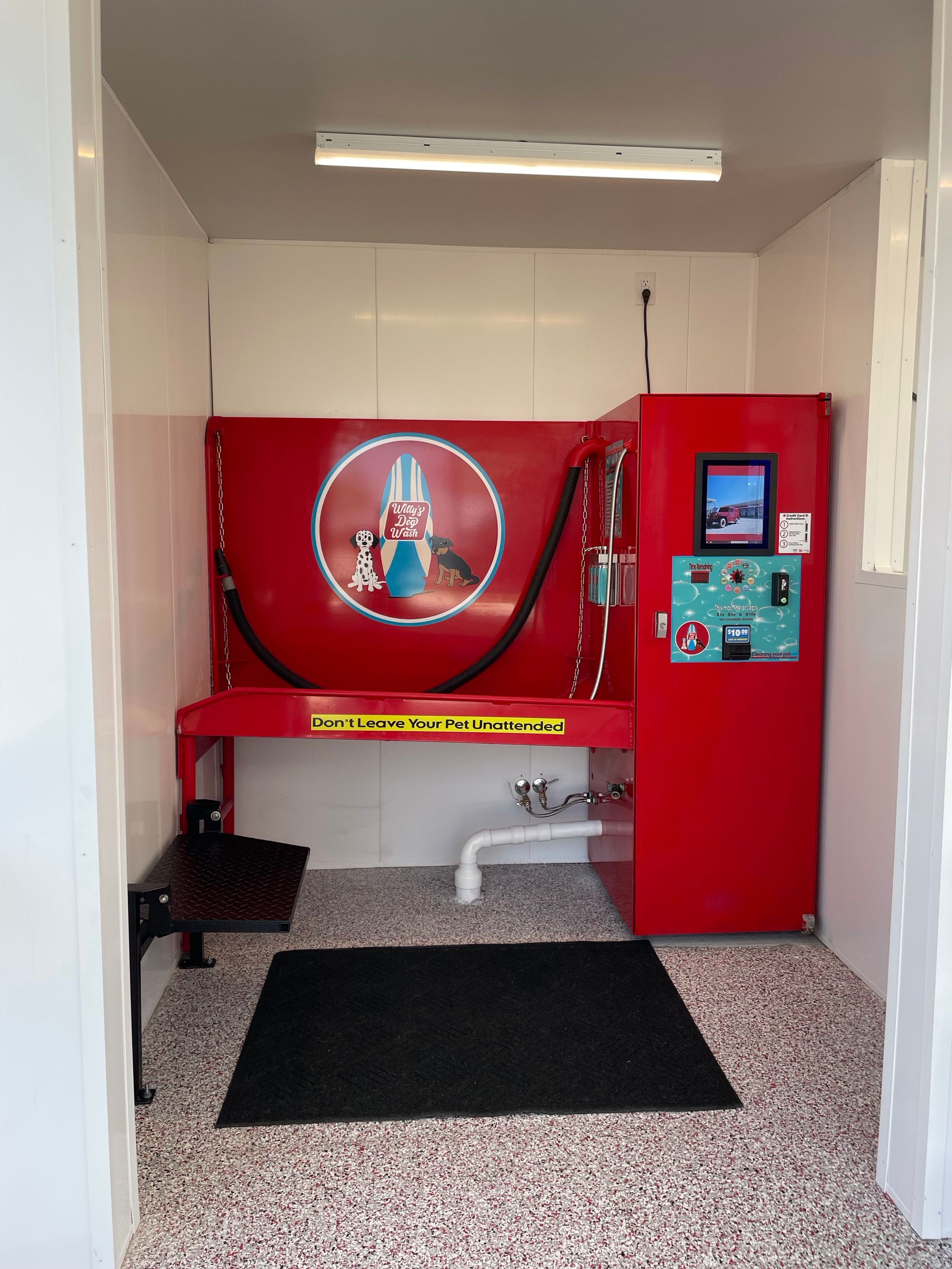 Small red arcade-style booth with a large emblem, keypad screen, and black floor mats in a hallway