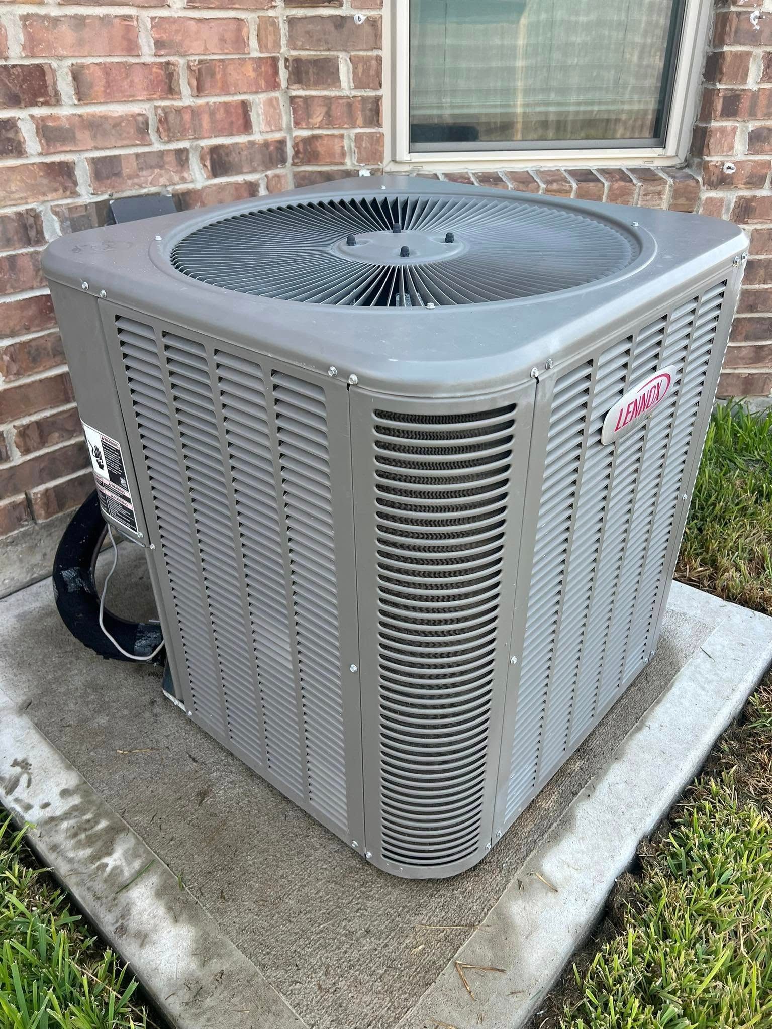 Outdoor air conditioning unit beside a brick house wall on a concrete pad.