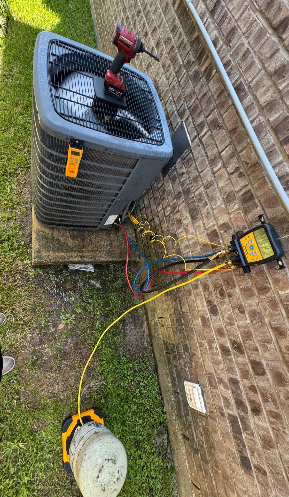 Outdoor HVAC unit with wires and tools on grass beside a brick wall, viewed from above