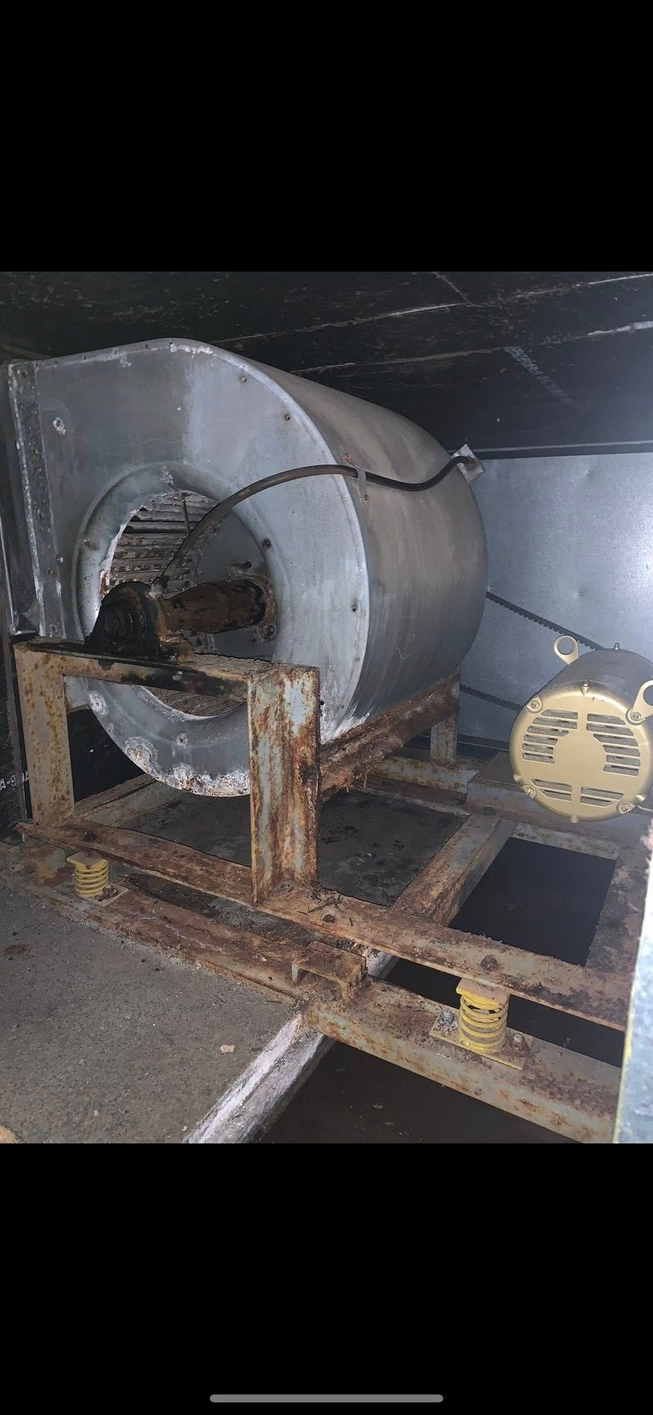 Large industrial fan with rusted frame and ductwork in a dark, dusty storage area