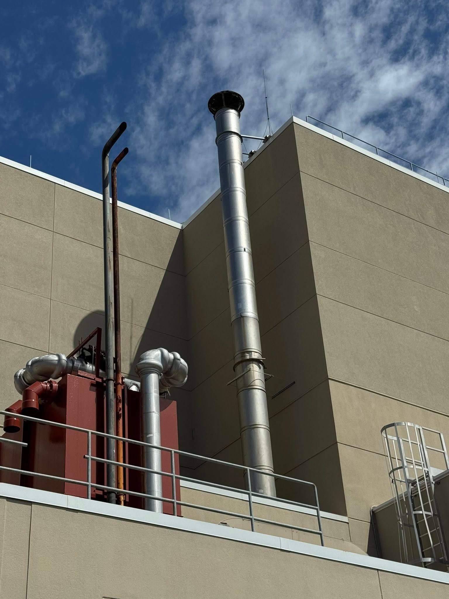 Industrial rooftop with silver vent pipe and red equipment against a beige building and blue sky
