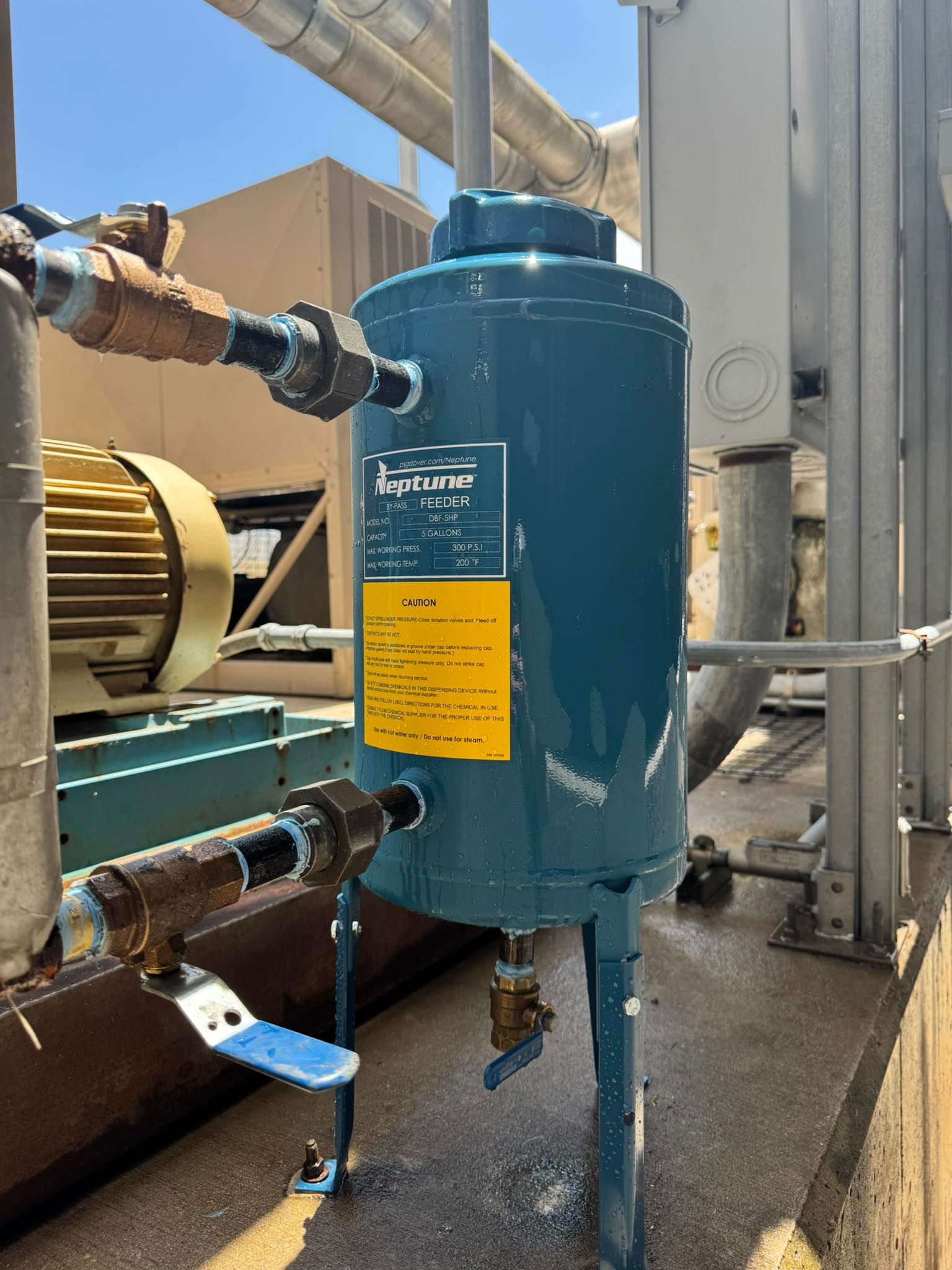 Blue industrial tank with pipes and yellow label beside machinery in an outdoor facility