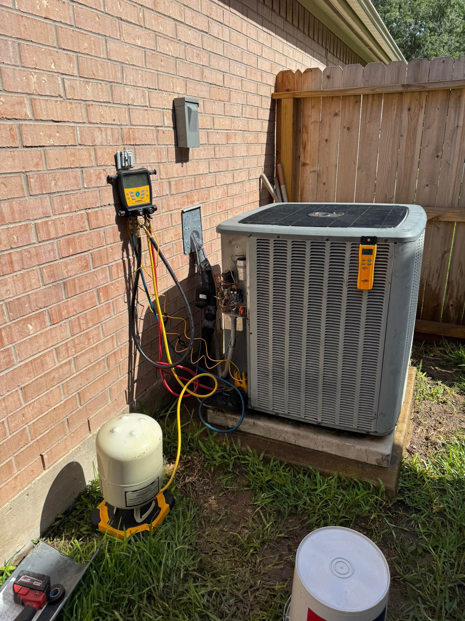 Outdoor HVAC unit and equipment beside a brick house, with hoses and control boxes in a fenced yard.