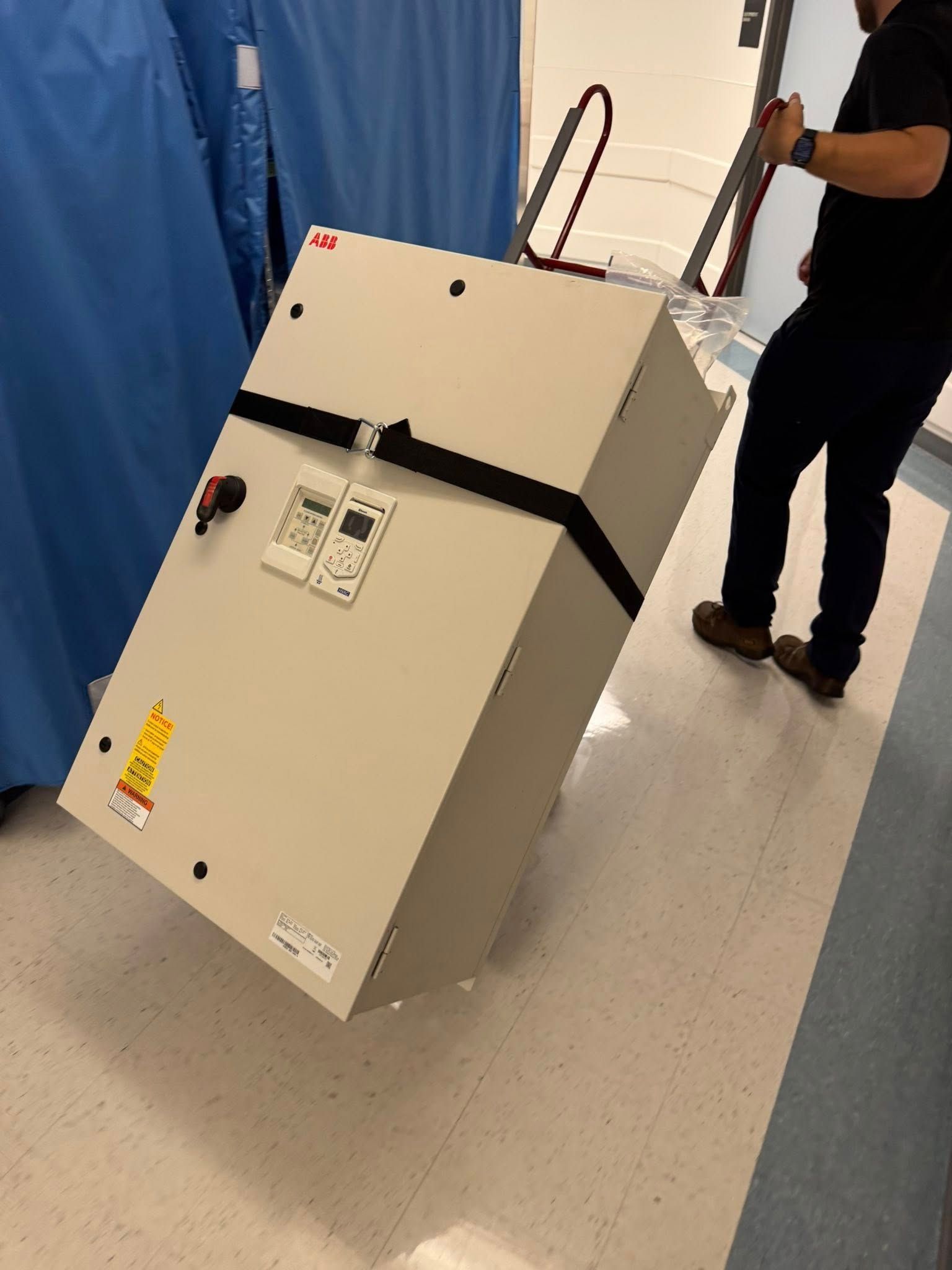 Large beige equipment cabinet on a wheeled dolly in a hallway, with a person holding it upright.