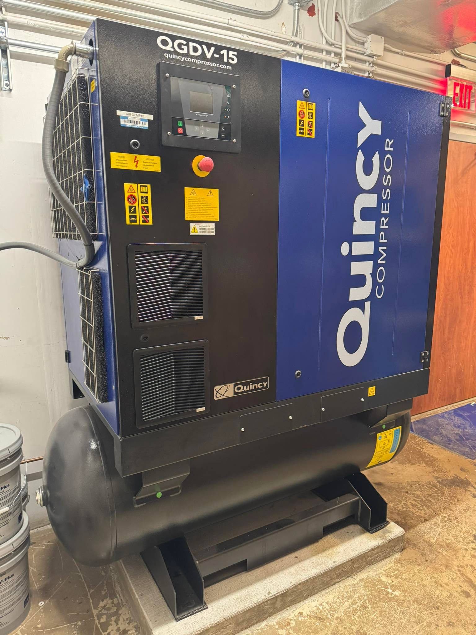 Blue Quincy air compressor in a workshop, with hoses and vents visible on a concrete floor.