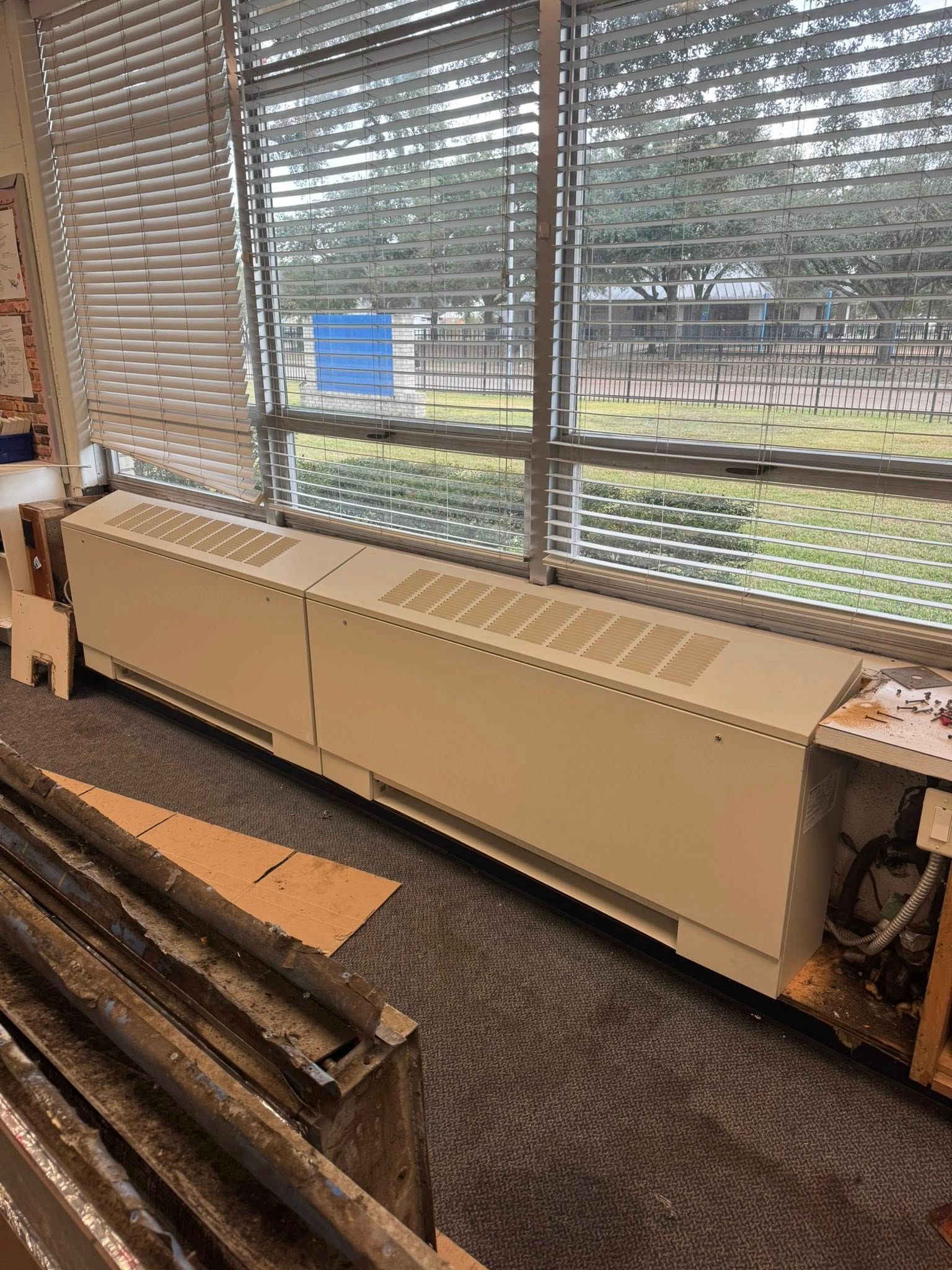 Long beige radiator under windows with blinds in an office or classroom, overlooking a grassy field