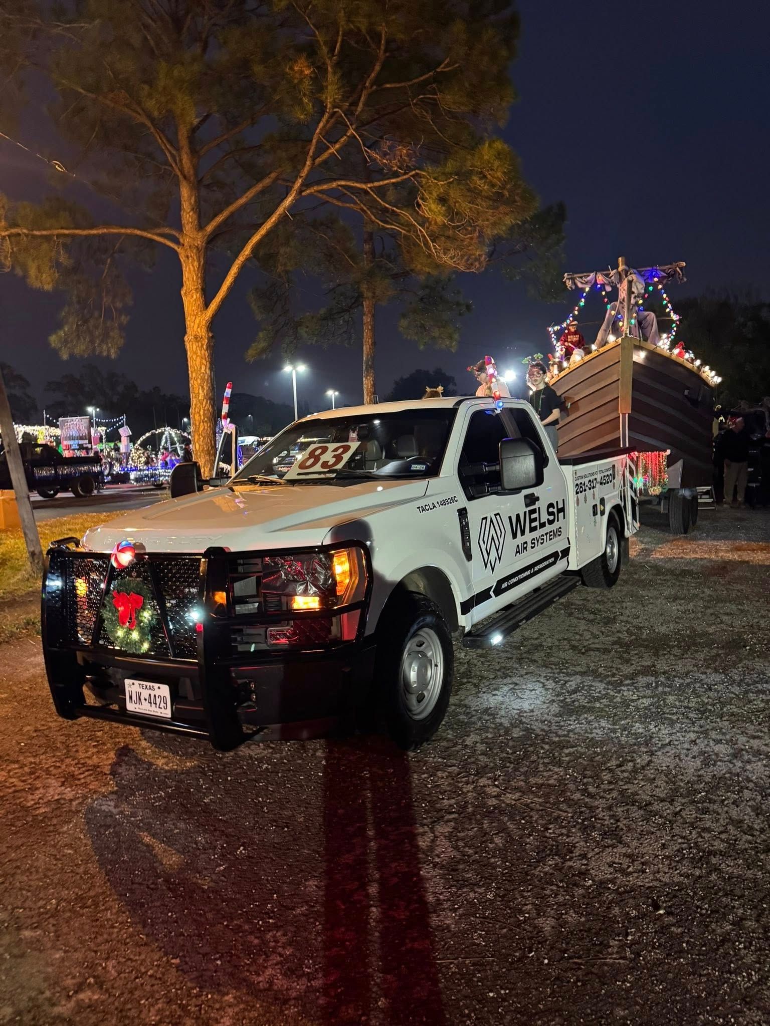 White tow truck decorated with Christmas lights at a nighttime parade, with a lit float behind it.